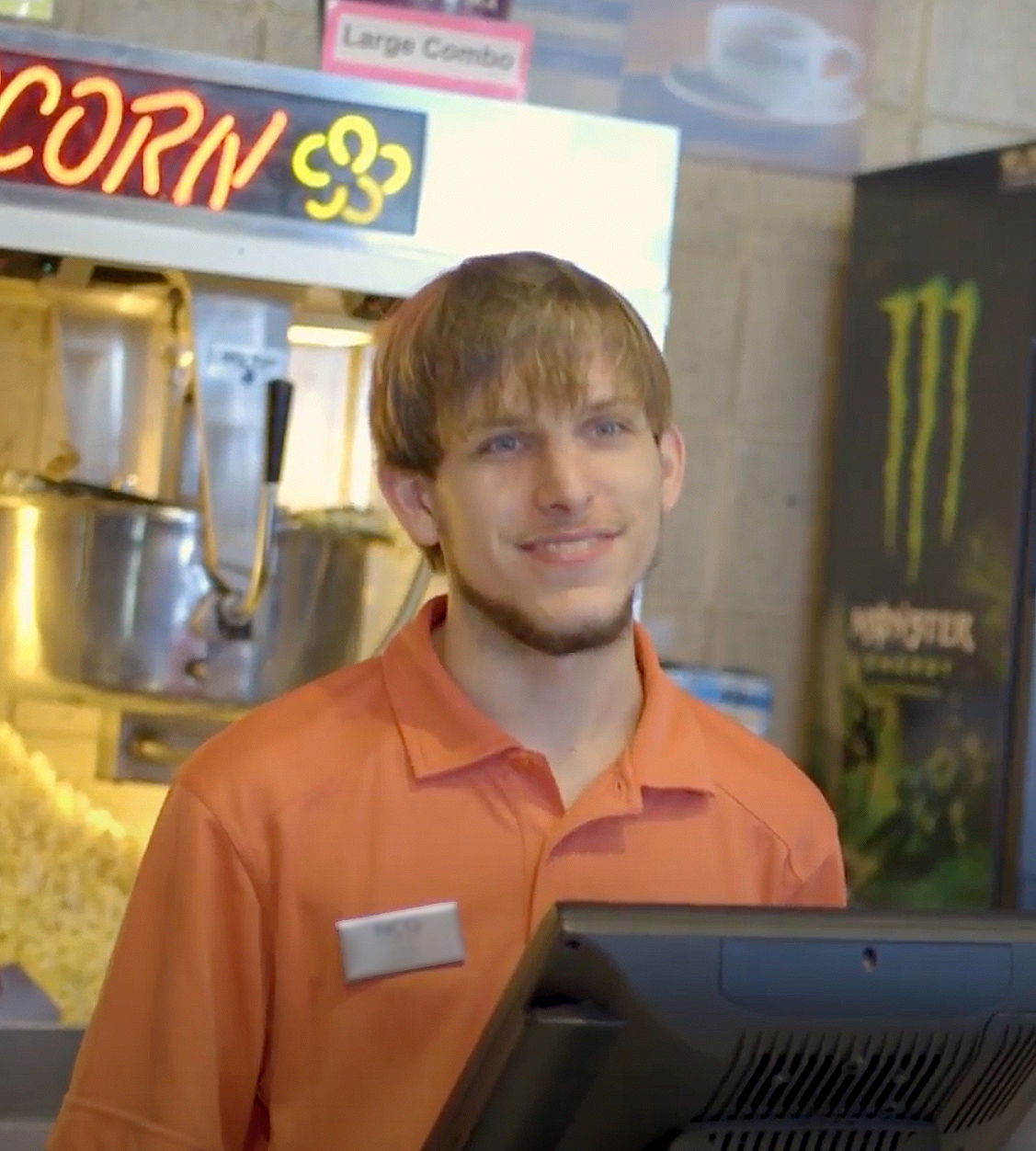 employee in orange polo standing in front of a popcorn maker and behind a cash register